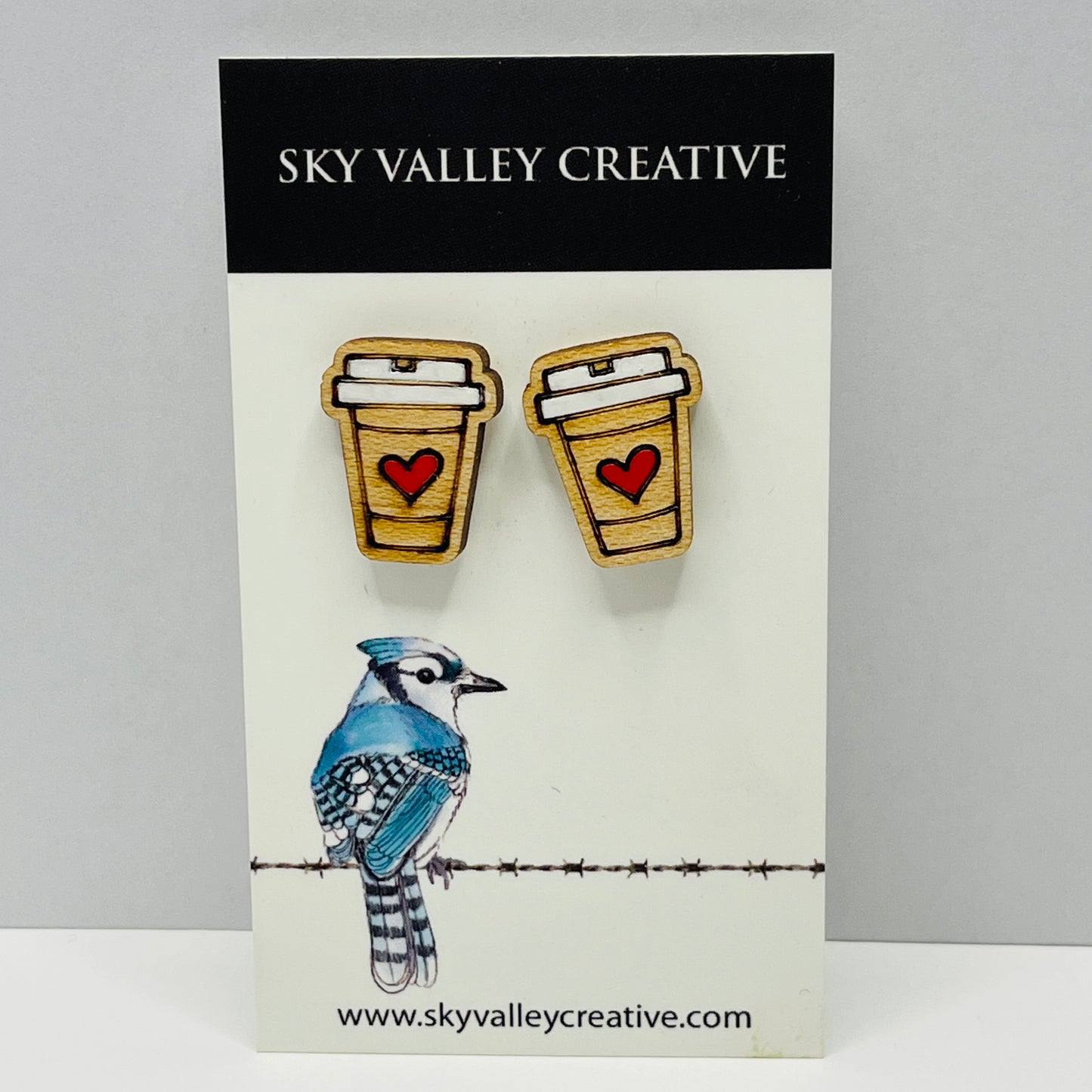 Pair of earrings shaped like coffee cups with a heart on a card featuring a blue jay illustration, branded 'Sky Valley Creative'.