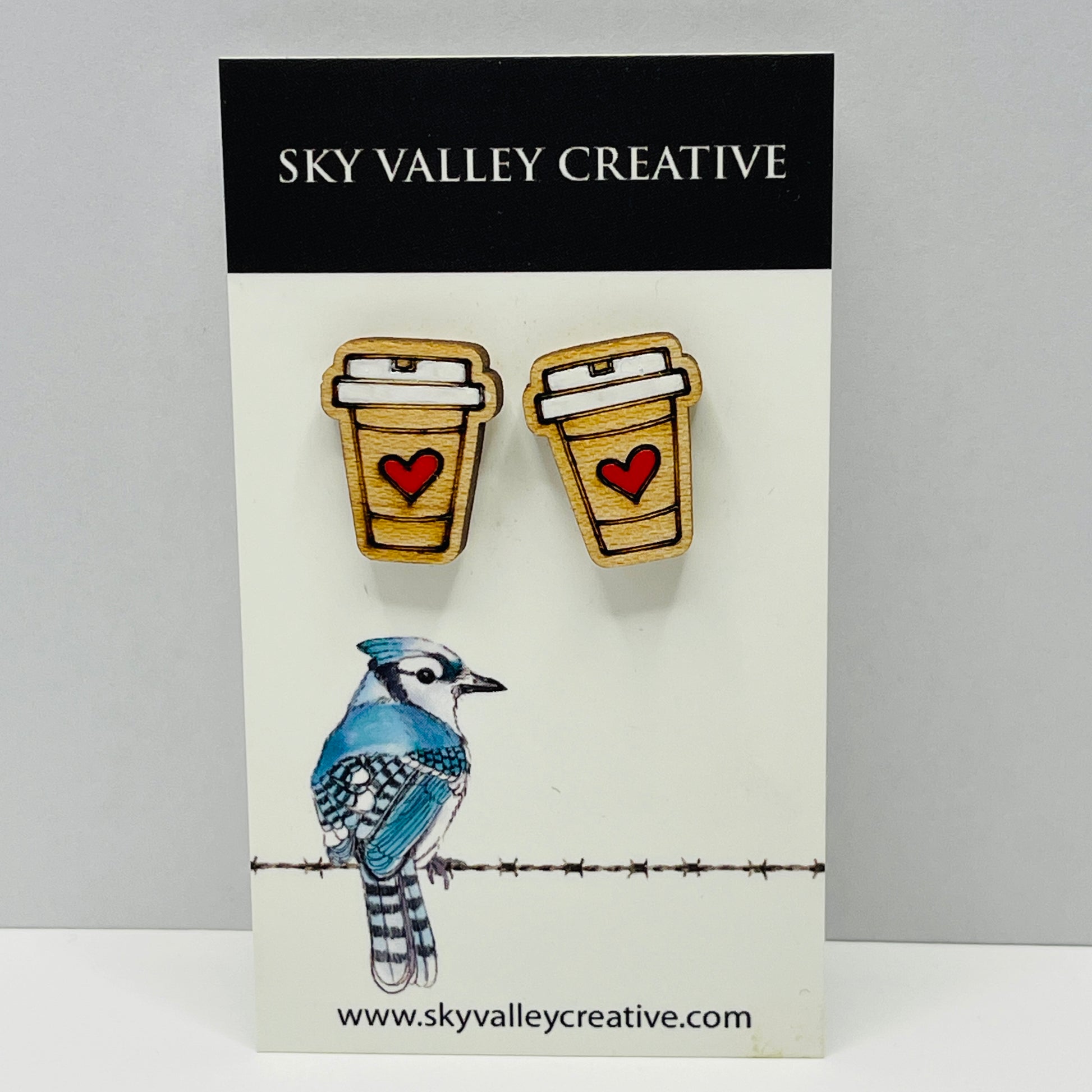 Pair of earrings shaped like coffee cups with a heart on a card featuring a blue jay illustration, branded 'Sky Valley Creative'.