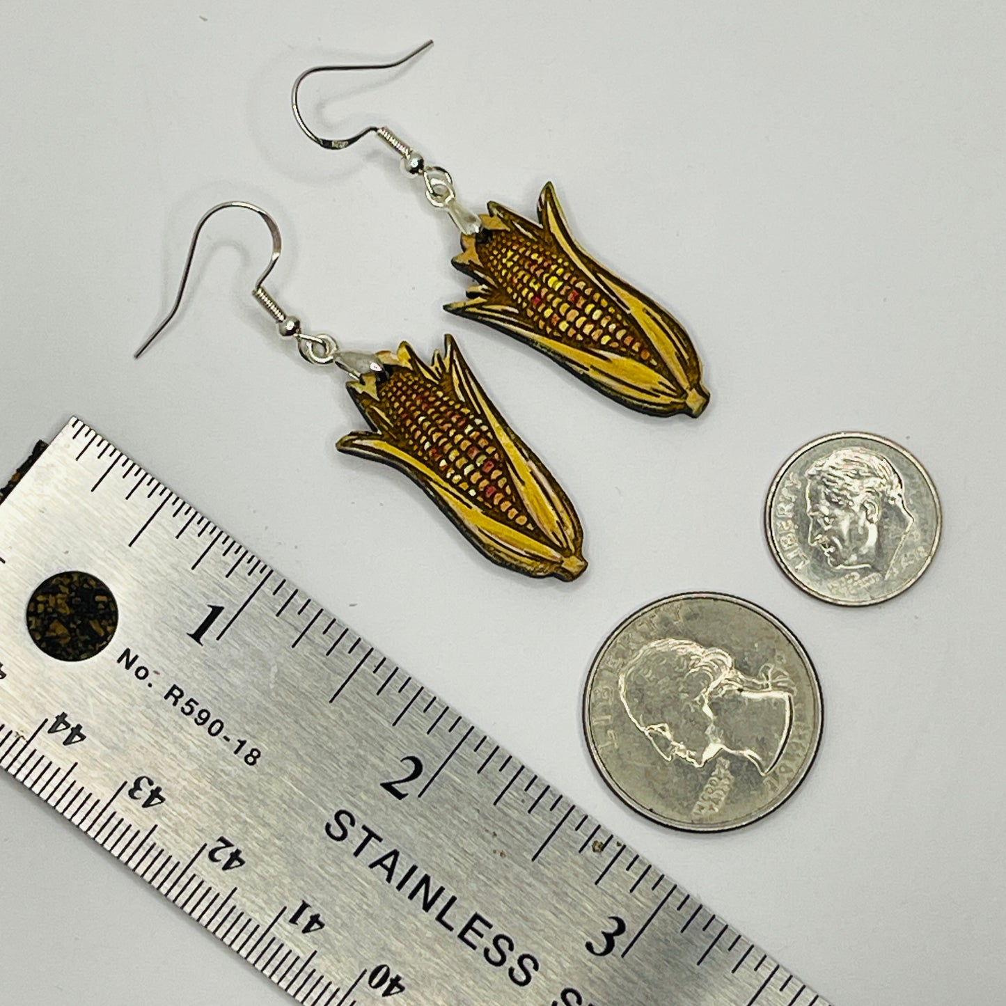 Corn-shaped earrings next to a ruler and coins (for scale) on a white background