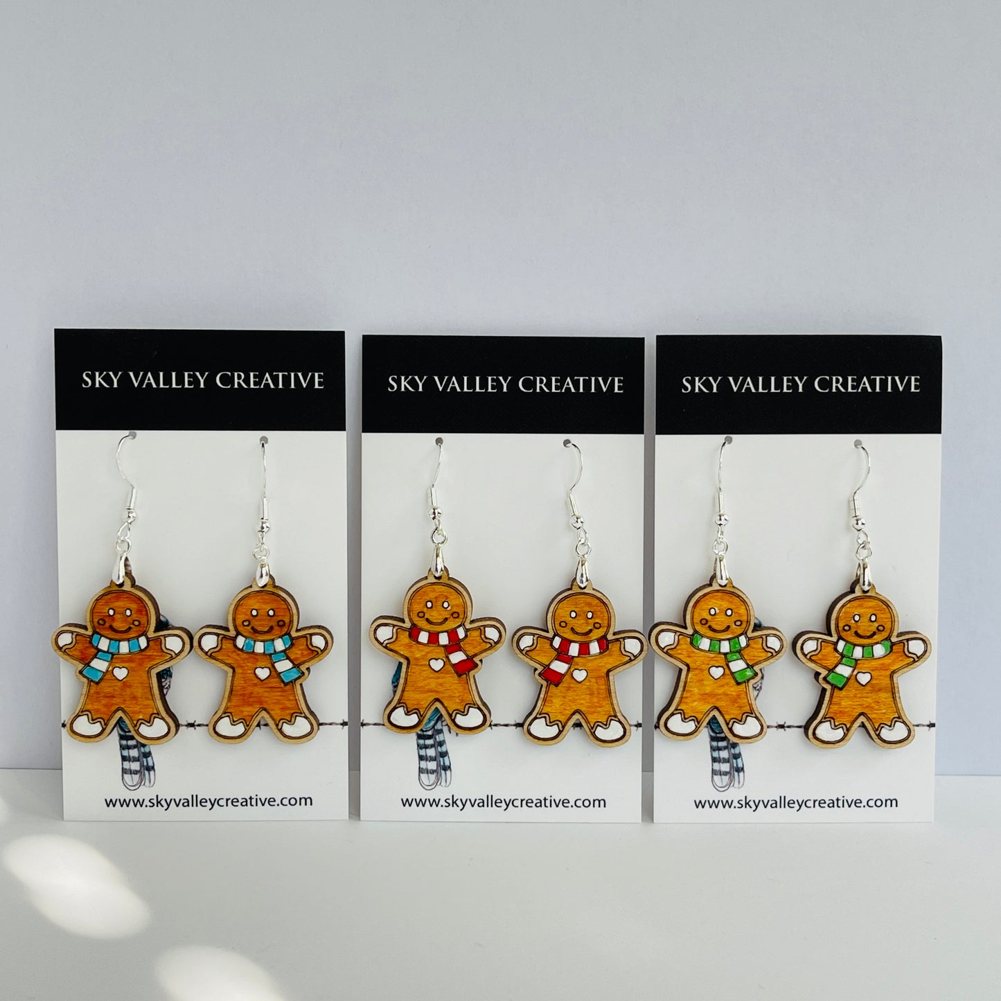 Three pairs of gingerbread man earrings with blue, red and green scarf choices on display cards with 'Sky Valley Creative' branding.